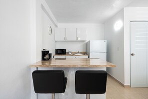 Una cocina compacta con armarios blancos, microondas, cafetera y refrigerador. Hay dos sillas negras en la barra.