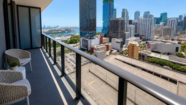 Uma varanda com cadeiras de vime, vista para a paisagem urbana incluindo um prédio alto em construção e um estacionamento.