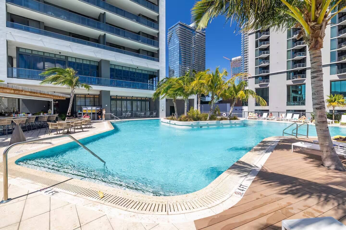 A swimming pool with lounge chairs and palm trees, surrounded by modern buildings.