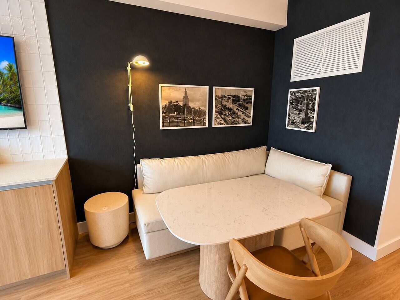 Una habitación moderna con una mesa redonda, sofá color beige y un taburete pequeño. Hay tres cuadros enmarcados en la pared, una lámpara y una ventana con persianas.