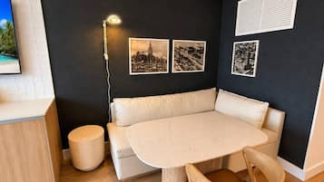 A modern room with a round table, beige sofa, and a small stool. There are three framed pictures on the wall, a lamp, and a window with blinds.
