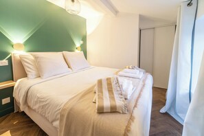 En snyggt bäddad säng med vita sängkläder och en beige pläd, en väggmonterad lampa och ett trägolv.