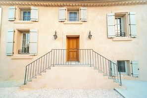 Un edificio con una puerta central de madera, varias ventanas con contraventanas y una escalera que conduce a la entrada.