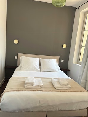 A neatly made bed with pillows and folded towels, a bedside table, and a wall-mounted light fixture. - Garrigue house at the heart of a vineyard estate (Bages)
