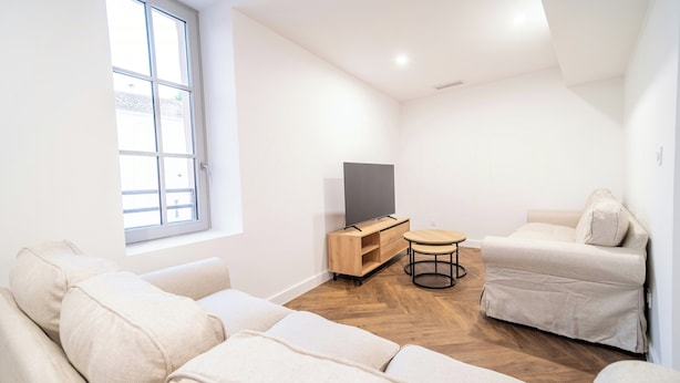 Ein modernes Wohnzimmer mit einer Sofagarnitur, einem Fernseher auf einem Holzboden, einem kleinen runden Tisch und einem Fenster mit Blick nach außen.
