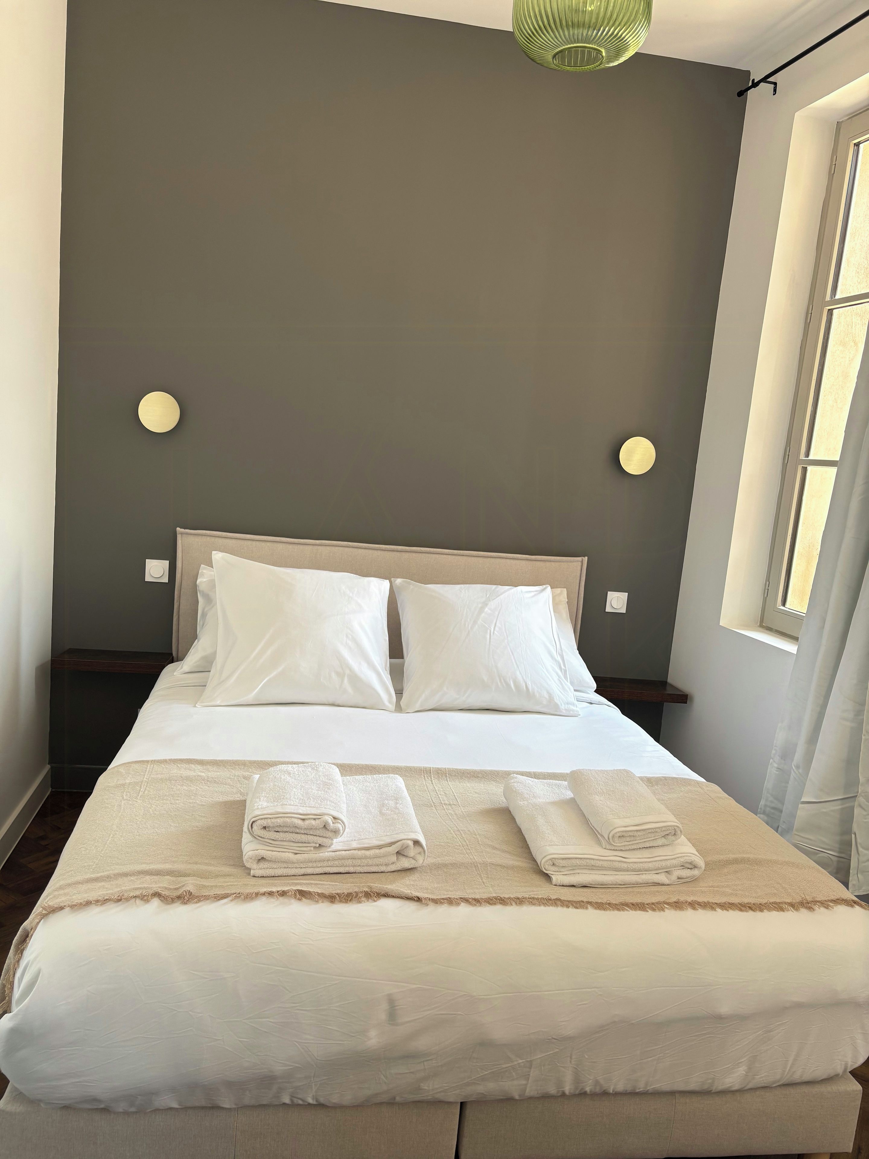 A neatly made bed with pillows and folded towels, a bedside table, and a wall-mounted light fixture.