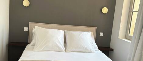 A neatly made bed with pillows and folded towels, a bedside table, and a wall-mounted light fixture.
