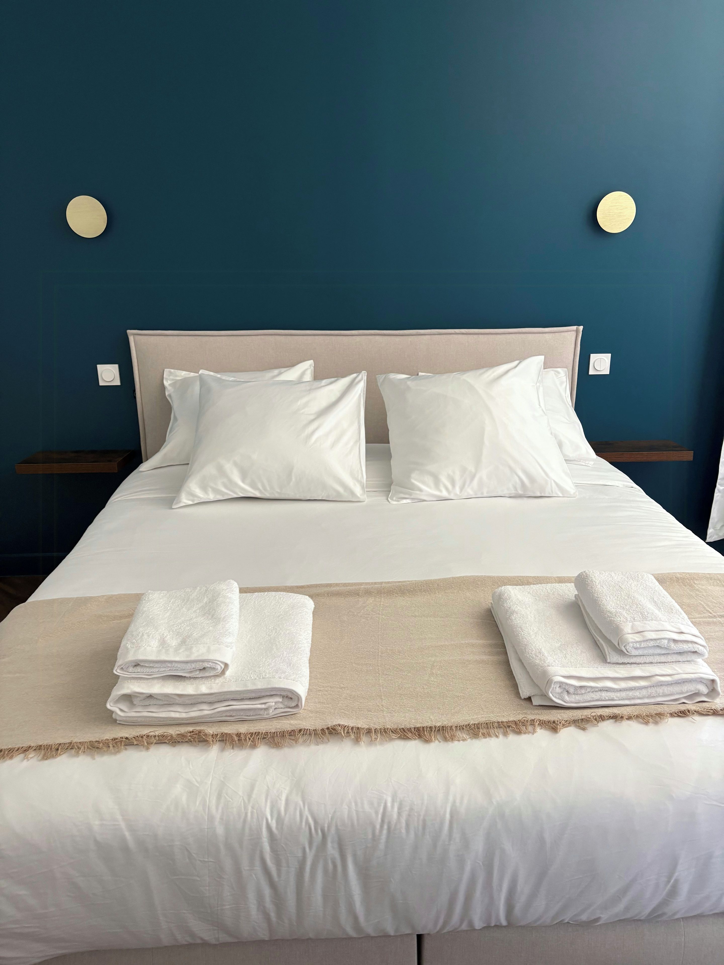 A neatly made bed with white linens, two pillows, and two folded towels on a beige bedspread.