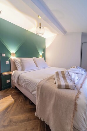 A neatly made bed with white linens and a beige throw blanket, positioned in a room with teal walls and wooden flooring. - Garrigue house at the heart of a vineyard estate (Bages)