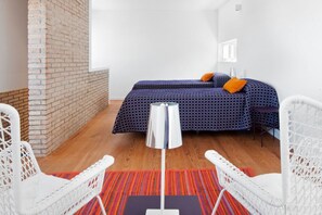 Une pièce avec un mur en briques, un lit recouvert d’une literie à motifs, un lampadaire et des chaises en osier peintes en blanc.