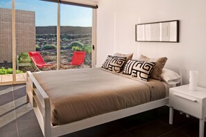 A bedroom with a bed, a nightstand, a window with a view of the desert, and a red chair. - Casas Bioclimáticas Iter El Alisio (Granadilla de Abona)