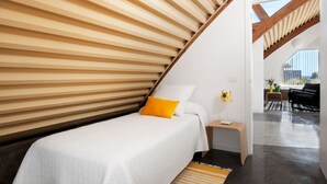 A bedroom with a sloped ceiling, a bed with white bedding and a yellow pillow, a bedside table with a lamp, and a view of the outdoors. - Casas Bioclimáticas Iter El Cangrejo (Granadilla de Abona)