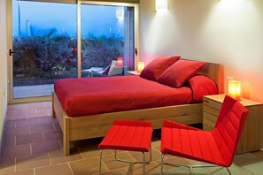 Ein Schlafzimmer mit einem Bett, zwei roten Sesseln, einem Nachttisch und Blick auf einen Garten durch eine Schiebetür.