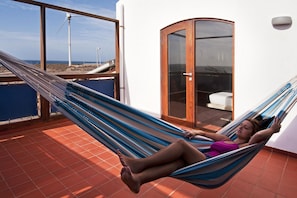 A person lying in a hammock on a balcony with a view of wind turbines and a beach. - Casas Bioclimáticas Iter La Arcilla (Granadilla de Abona)