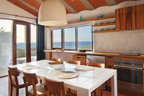 A modern kitchen with a view of the sea, featuring a white table, wooden chairs, and a hanging light fixture. - Casas Bioclimáticas Iter La Arcilla (Granadilla de Abona)