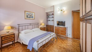 Quarto com cama, mesas de cabeceira, televisão, cômoda e estante.