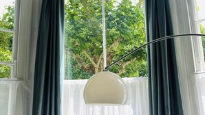 Quarto moderno com mesa de vidro redonda, duas cadeiras brancas, uma planta em vaso e uma janela grande com cortinas.