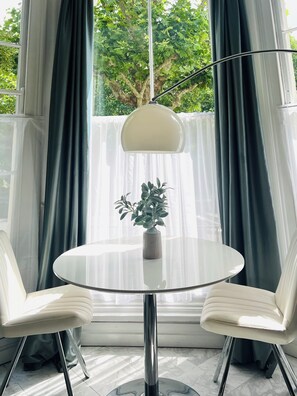 A modern room with a round glass table, two white chairs, a potted plant, and a large window with curtains. - Luxury flat in Notting Hill, 4 mins Portobello Market. 3 large double beds. (London)