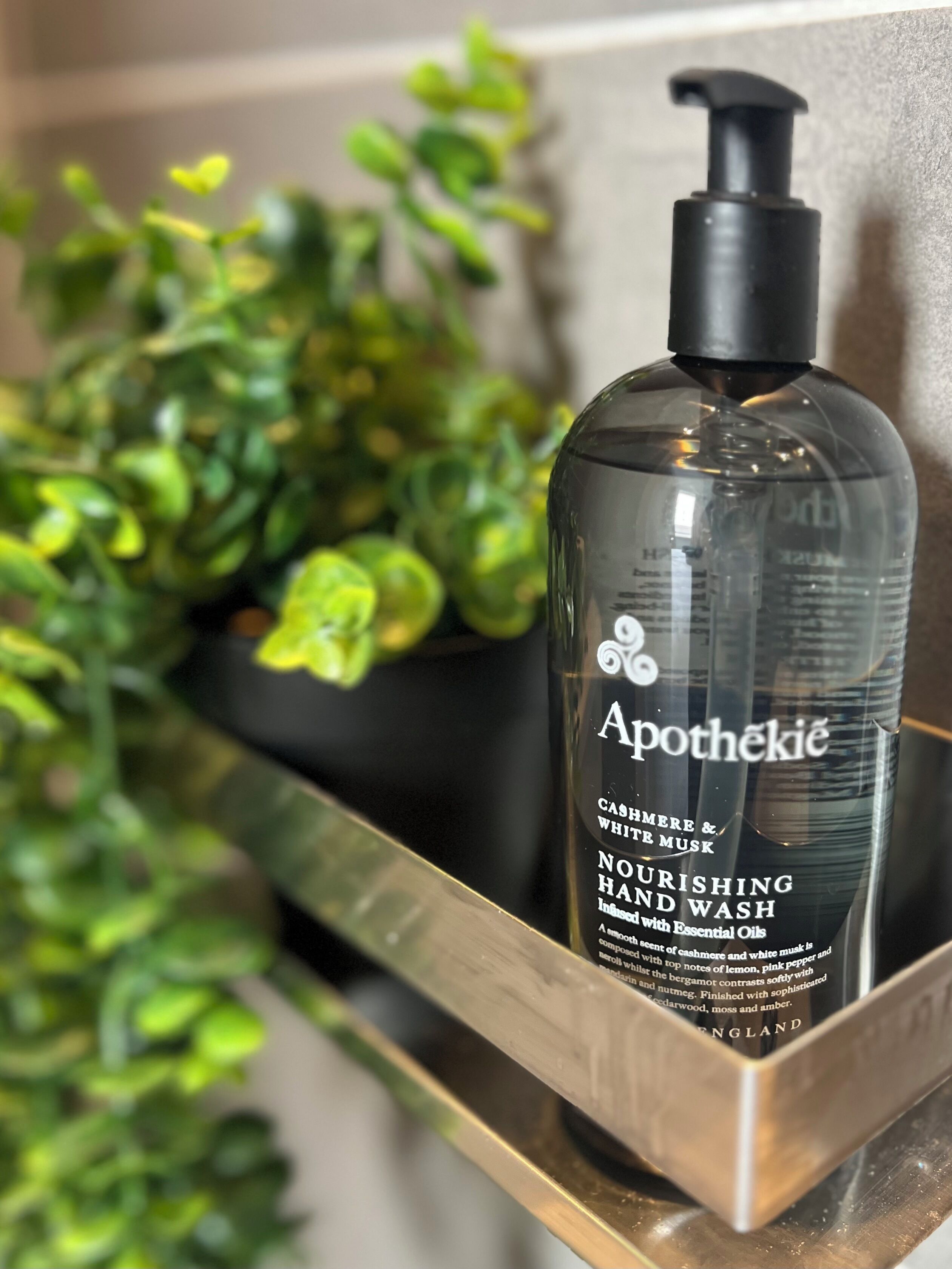 Une bouteille de savon pour les mains de la marque Apothéki est posée sur une étagère, avec une plante en pot en arrière-plan.