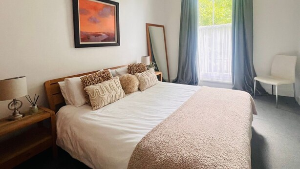 Quarto de hotel com cama, mesa de cabeceira com luminária, cadeira, janela com cortinas e quadro na parede.