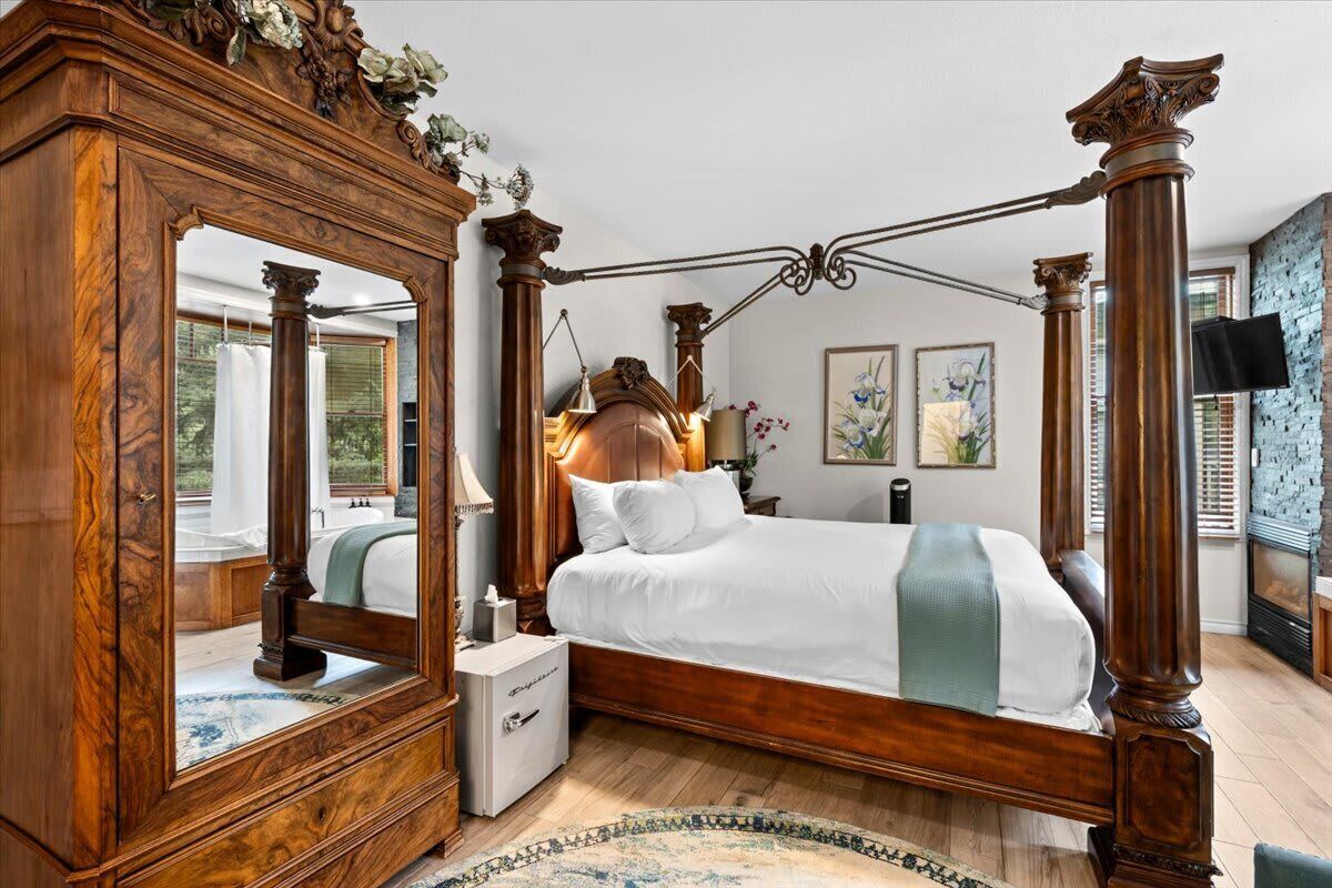 A four-poster bed with a wooden frame and white bedding, a large mirror, and a small bedside table with a lamp.