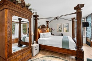 Ein Bett mit Baldachin, dessen Holzbettgestell mit weißer Bettwäsche bezogen ist, ein großer Spiegel und ein kleiner Nachttisch mit einer Lampe.