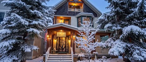 A two-story house with a snow-covered front yard, a wooden porch, and a visible American flag.
