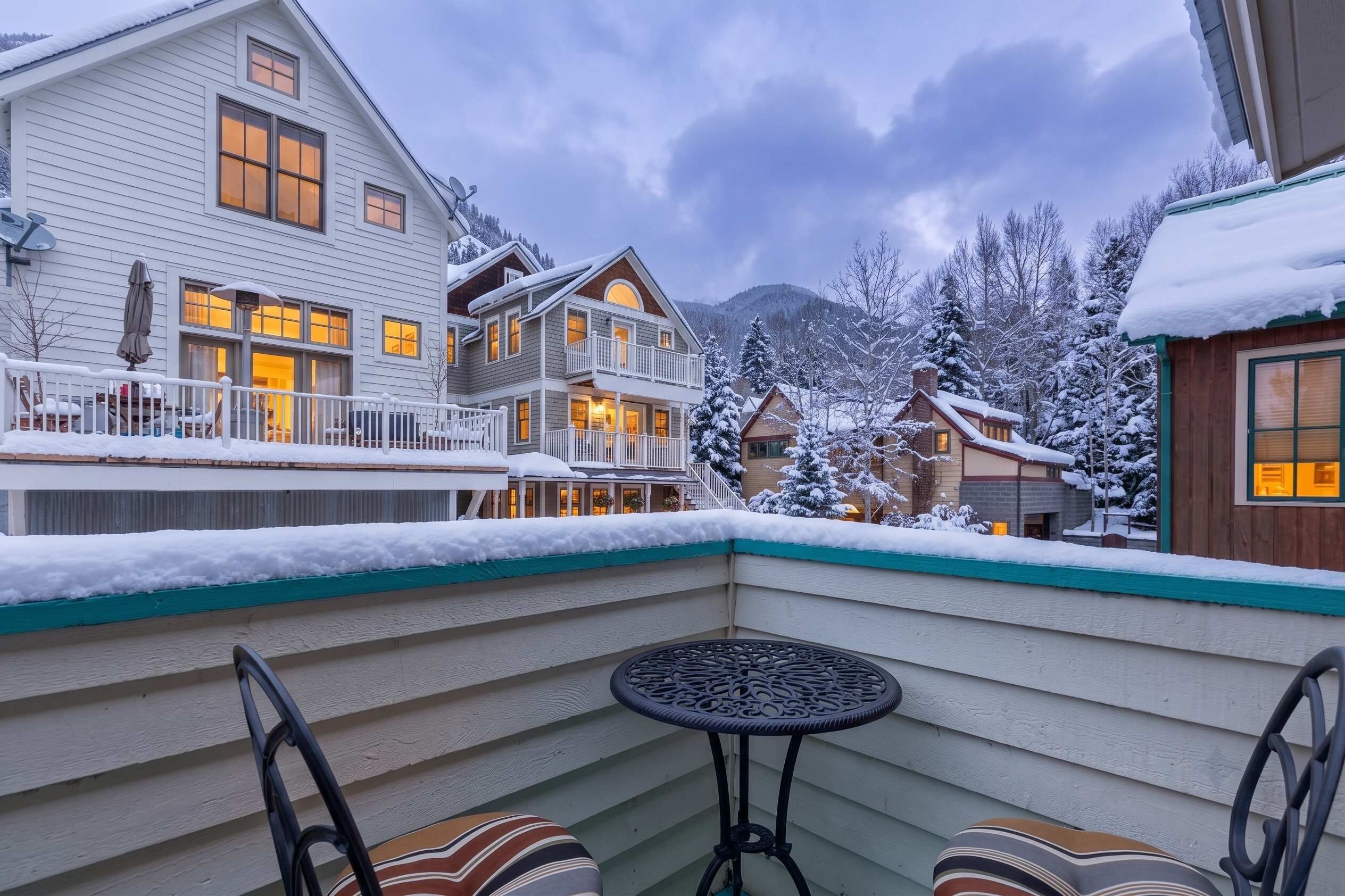 A snow-covered balcony with a table and chairs, overlooking a residential area with houses and a mountain backdrop.