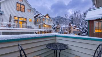 A snow-covered balcony with a table and chairs, overlooking a residential area with houses and a mountain backdrop.