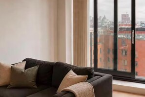 A dark-colored sofa with multiple cushions in a room with a large window overlooking a cityscape. - Salford Serenity - Modern 2 Bedroom Apartment (Salford)