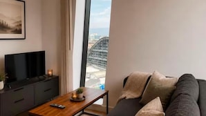A modern living room with a sofa, a coffee table, a TV, and a large window offering a view of a bridge. - Salford Serenity - Modern 2 Bedroom Apartment (Salford)