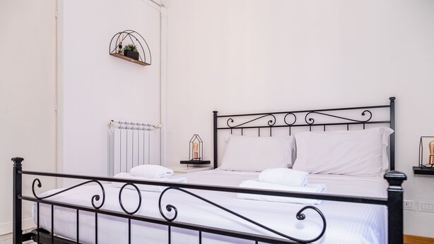 A neatly made bed with white linens, a black metal headboard, and a bedside table with a lamp. - Aurelia Vatican Home (Roma)