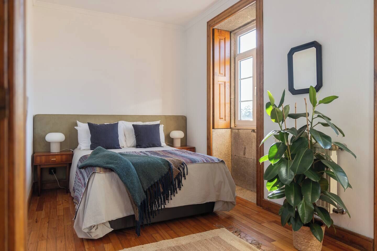 A bedroom with a bed, bedside tables, a large potted plant, and a window.