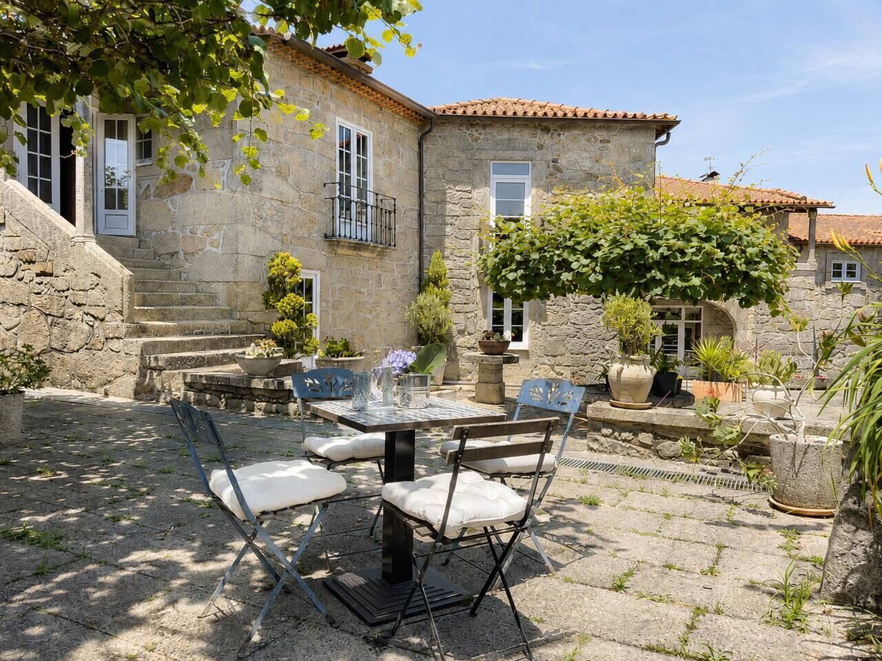 Ein Steinhaus mit einer Terrasse, auf der ein Tisch und Stühle stehen, umgeben von Topfpflanzen und einer Steintreppe.