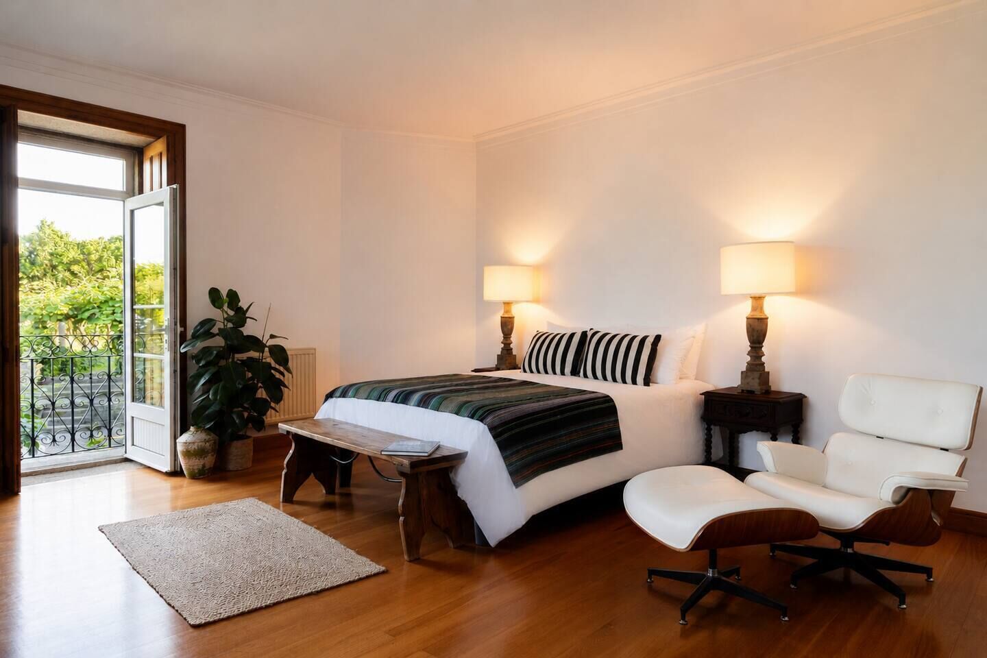 Ein Schlafzimmer mit Bett, Nachttischlampen, einem Sessel, einem Tisch, einer Zimmerpflanze und Blick durch eine Tür.