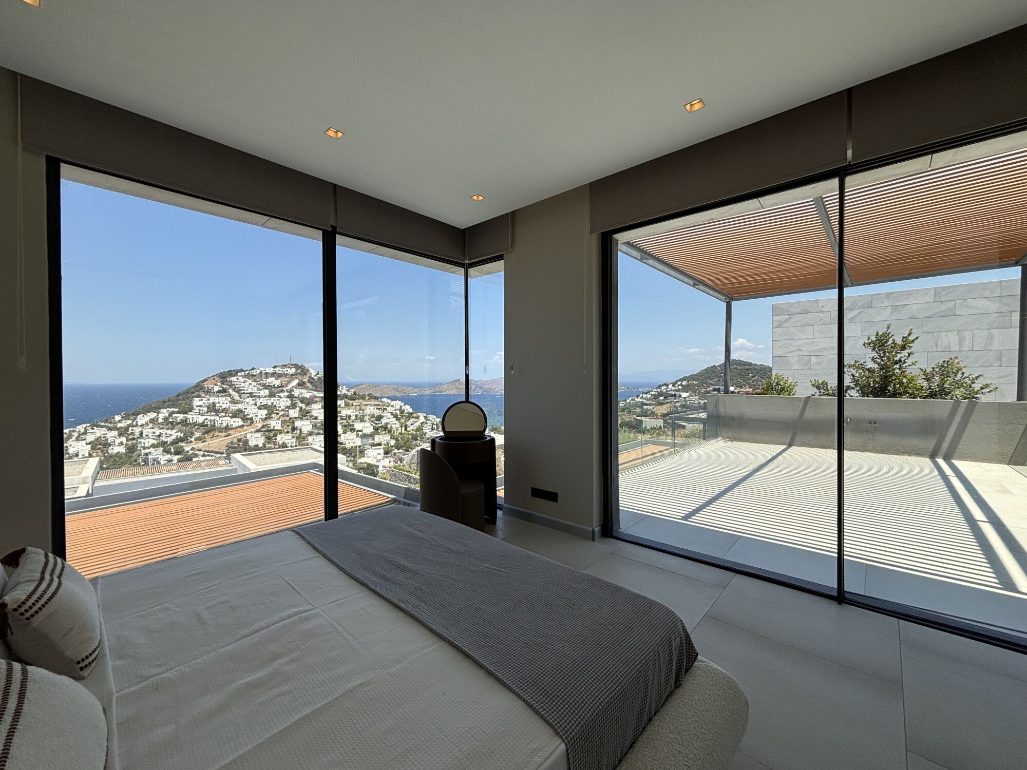 Une chambre moderne avec un grand lit, une petite table de chevet et une vue sur une ville côtière grâce  à de grandes fenêtres du sol au plafond.