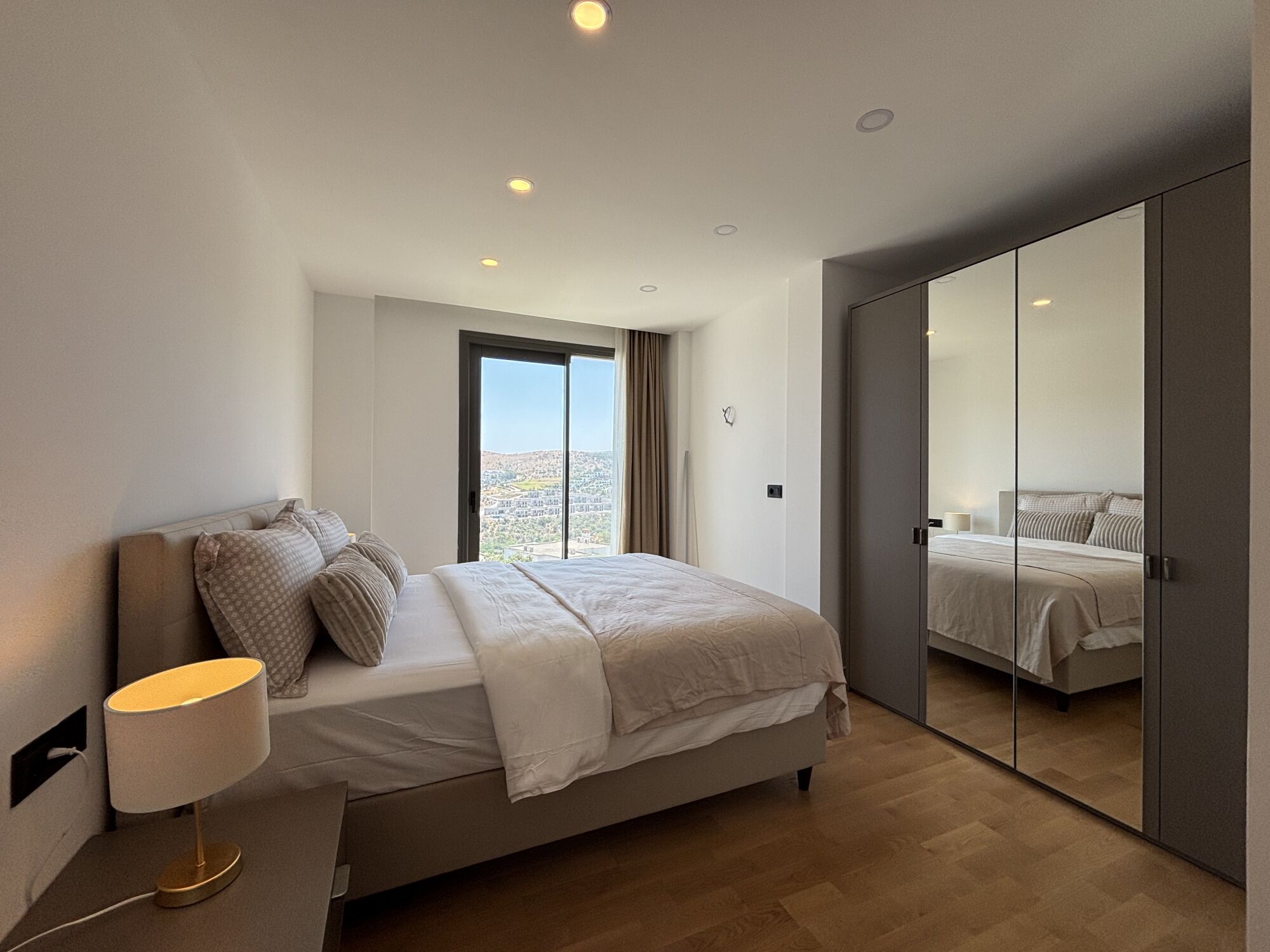 A modern bedroom with a bed, bedside lamp, and a view through a large window.