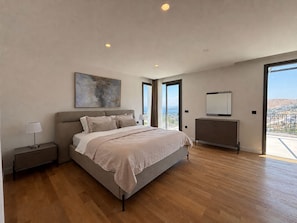 A bedroom with a large bed, a dresser, a nightstand, a painting, and a view of the outdoors.