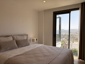 A bedroom with a large bed, a nightstand, and a view of a cityscape through a balcony.