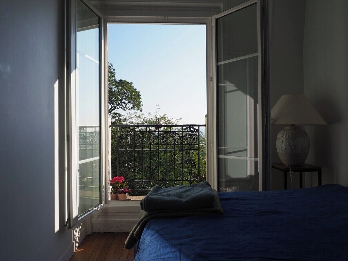 A bedroom with a large window, a bed, a nightstand, and a potted plant.