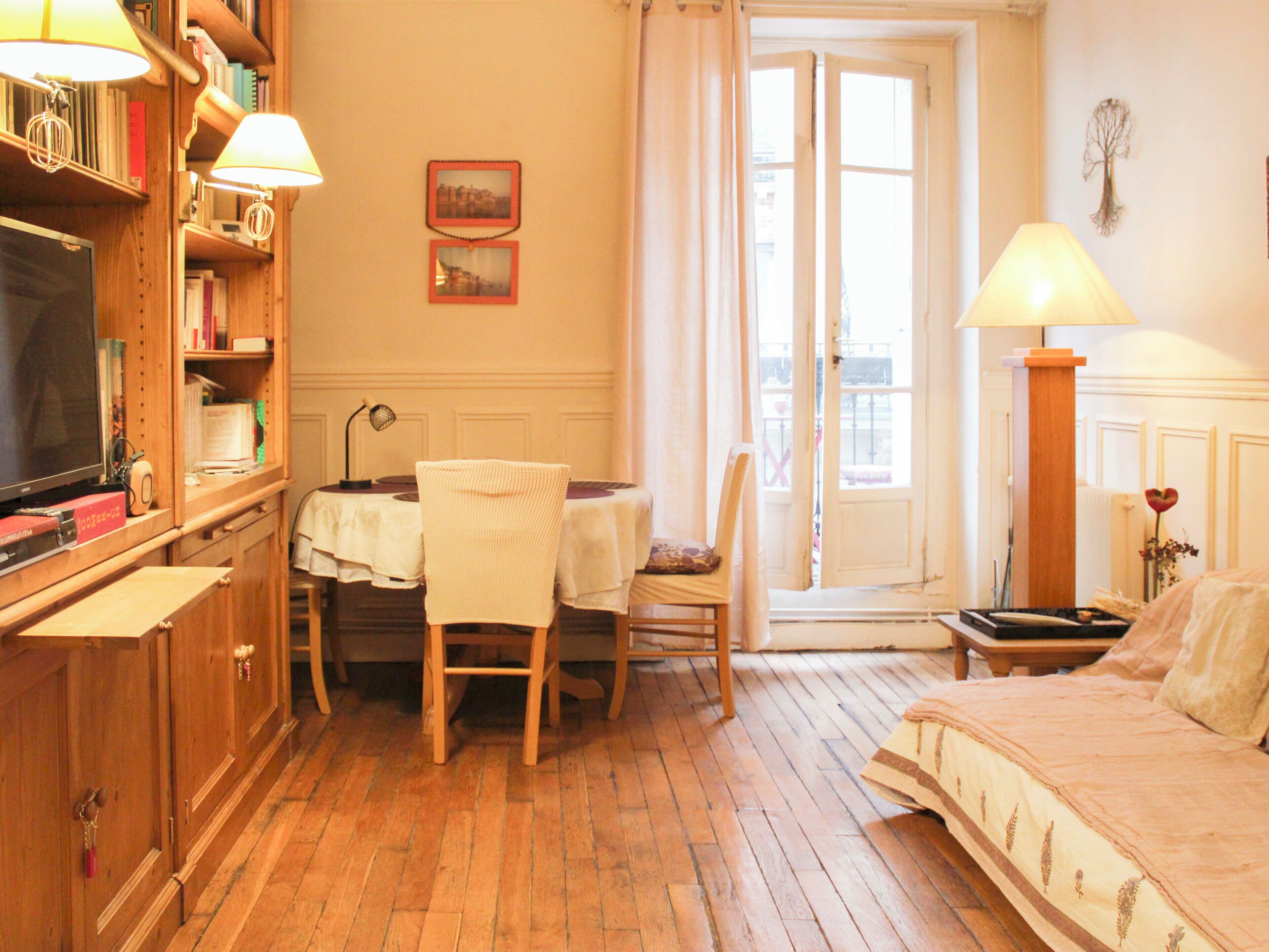 Une pièce avec un plancher en bois, un lit recouvert d’une couvre-lit à motifs floraux, une table avec des chaises, une bibliothèque, un téléviseur et une fenêtre avec des rideaux.