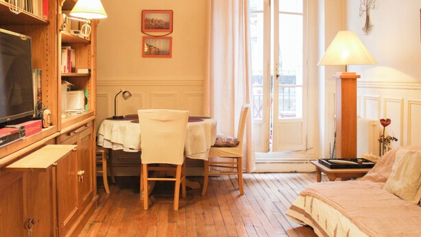 Una habitación con piso de madera, una cama con colcha estampada, una mesa con sillas, una estantería, un televisor y una ventana con cortinas.