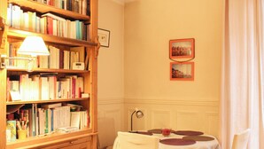 Una habitación con una estantería de madera llena de libros, una mesa con mantel blanco y sillas, y dos cuadros en la pared.