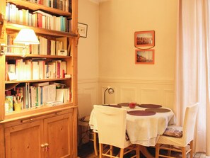 Ein Zimmer mit einer Holzhülle voller Bücher, einem Tisch mit weißem Tischleinentuch und Stühlen sowie zwei gerahmten Bildern an der Wand.