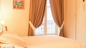 Una habitación con cama, ventana con cortinas, un cuadro en la pared y una mesita de noche con libros.