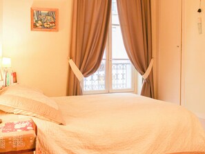Ein Zimmer mit einem Bett, einem Vorhangfenster, einem Bild an der Wand und einem Nachttisch mit Büchern.