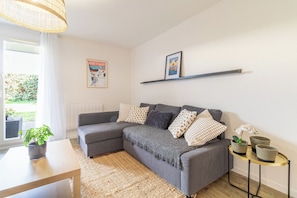 Ein Wohnzimmer mit einem grauen Ecksofa, einem kleinen runden Tisch, einem Blumentopf und einem Poster an der Wand.