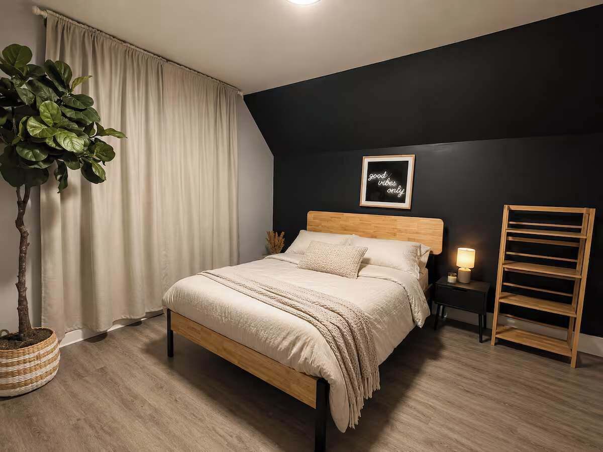 A bedroom with a bed, a bedside table with a lamp, a wall-mounted shelf, and a potted plant.