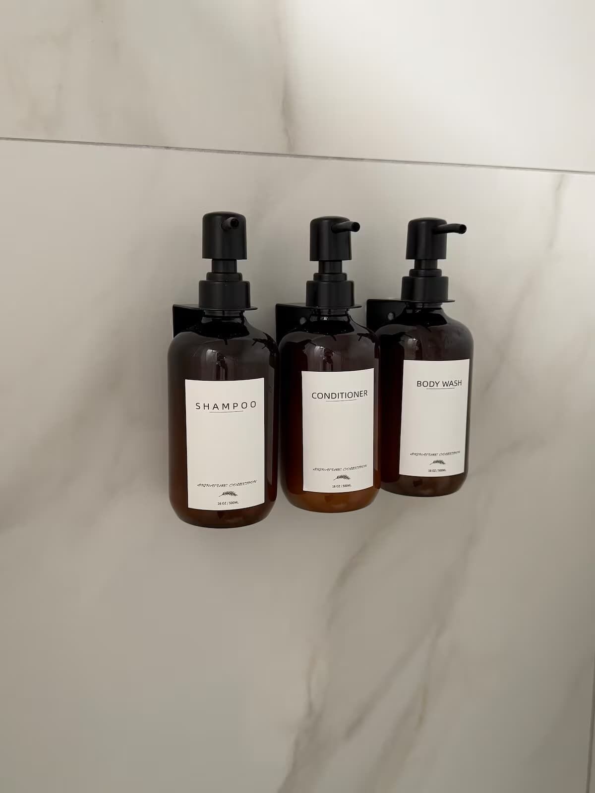 Three bottles of body wash and shampoo against a marble wall.