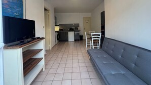 A living room with a grey sofa, a flat-screen TV on a stand, and a kitchen in the background. - Ground floor apart, walking distance to the beach (Cabourg)